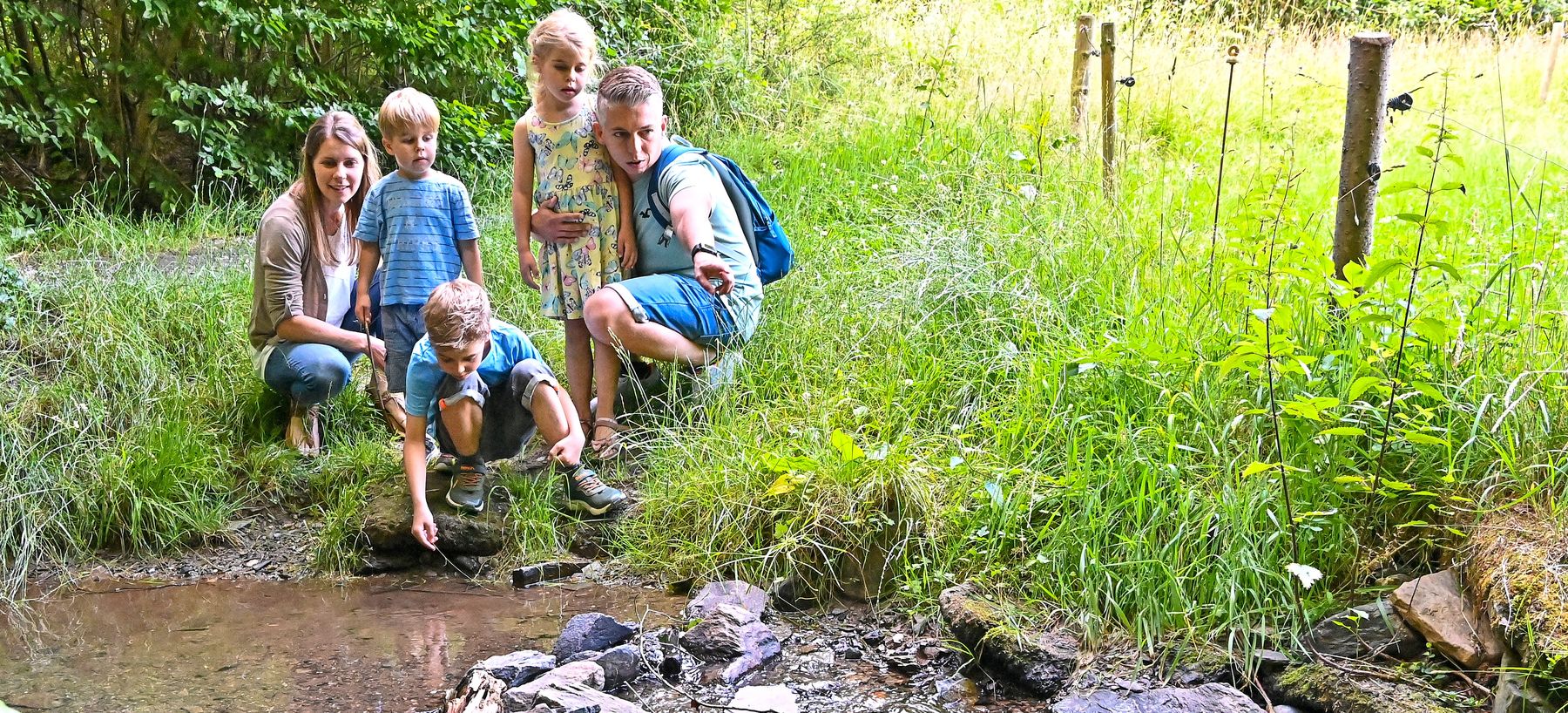 A woman, two boys, and two girls kneel and crouch by a small stream in a grassy, wooded area, looking at the water and exploring the natural surroundings together.